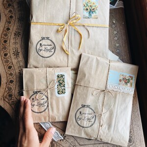 May include: Three parcels wrapped in brown paper, tied with twine and yellow ribbon. Each parcel has a sticker with a floral design or the text "Handmade by Leyla". The parcels are on a wooden surface.