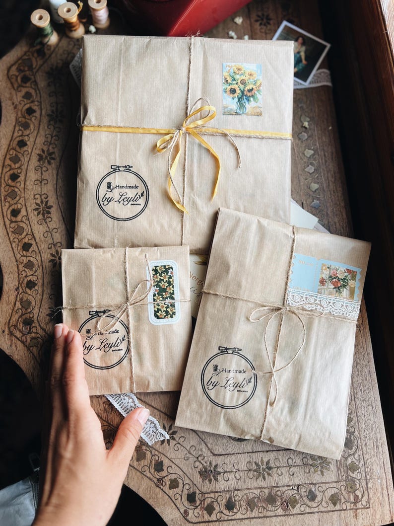 May include: Three parcels wrapped in brown paper, tied with twine and yellow ribbon. Each parcel has a sticker with a floral design or the text "Handmade by Leyla". The parcels are on a wooden surface.