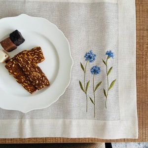 May include: A white linen placemat with a blue embroidered floral design. The placemat is on a wooden table with a white plate and cookies on it.