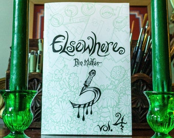 May include: A green book titled "Elsewhere" with the subtitle "Pin Mater" and the text "Vol. 24" on the cover. The book is sitting on a wooden table between two green candlesticks.