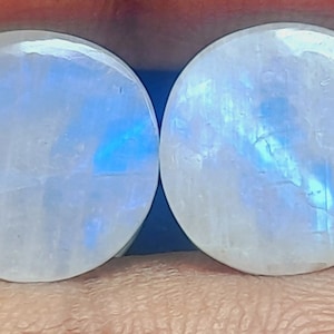 Könnte beinhalten: Zwei runde, weiße Mondstein-Cabochons mit einem blauen, irisierenden Schimmer.