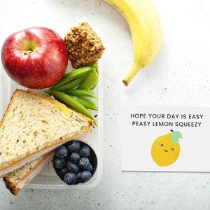 May include: A clear plastic lunch container filled with a sandwich, red apple, blueberries, snap peas, and a baked treat. A yellow banana and a card with a smiling lemon and the text "Hope your day is easy peasy lemon squeezy" are also present.