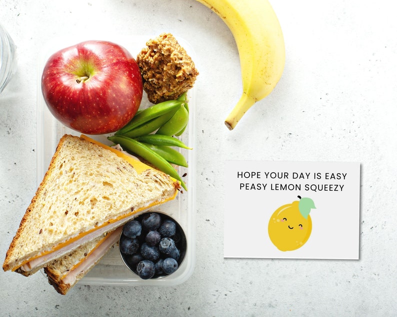 May include: A clear plastic lunch container filled with a sandwich, red apple, blueberries, snap peas, and a baked treat. A yellow banana and a card with a smiling lemon and the text "Hope your day is easy peasy lemon squeezy" are also present.