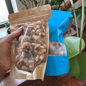 Op de afbeelding: Twee stazakken met lichtbruine, met suiker omhulde noten. De ene zak is gemaakt van natuurlijk kraftpapier met een doorzichtig plastic venster, terwijl de andere een felblauwe zak is met een rechthoekig doorzichtig venster in het midden.