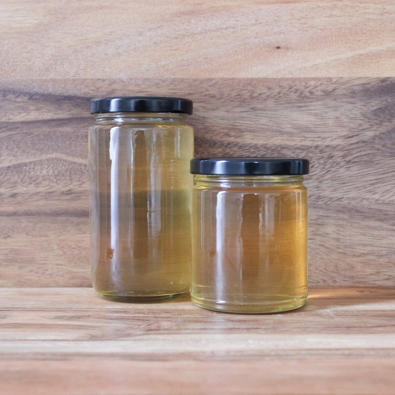 May include: Two glass jars with black lids filled with a light yellow liquid. The jars are sitting on a wooden surface.