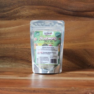 Pode incluir: Um saco transparente de chá de especiarias de noz-moscada de Granada está sobre uma superfície de madeira. A embalagem apresenta o texto "Nutmeg Spice Tea" e lista ingredientes como noz-moscada, canela, gengibre, açafrão e folha de louro. A marca "GCNA Grenada" também é visível.