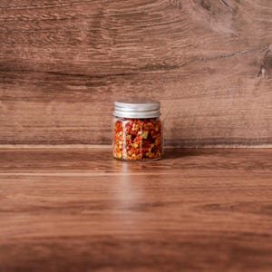 May include: A small, clear glass jar with a silver lid filled with red pepper flakes. The jar is centered on a wooden surface with a wooden background. The flakes appear to be a mix of red and orange.