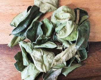 May include: A pile of dried kaffir lime leaves on a wooden surface. The leaves are green and brown, and some have a slightly curled shape.