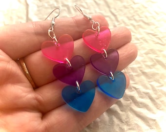 May include: A pair of dangle earrings with three layered heart charms. The hearts are pink, purple, and blue, and are made of clear acrylic. The earrings are silver.