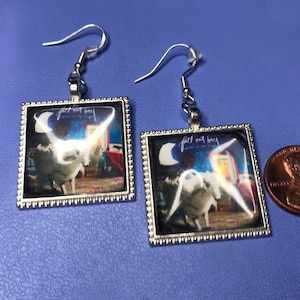 May include: A pair of silver earrings with a square glass charm featuring the Fall Out Boy album cover for "Save Rock and Roll". The album cover features a white unicorn with a blue background and a crescent moon.