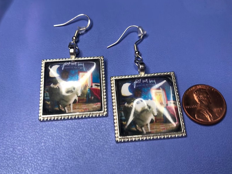 May include: A pair of silver earrings with a square glass charm featuring the Fall Out Boy album cover for "Save Rock and Roll". The album cover features a white unicorn with a blue background and a crescent moon.