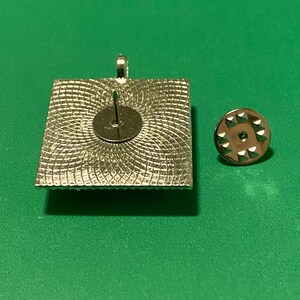 May include: The back of a silver square pin with a decorative pattern. The pin has a silver pin back.