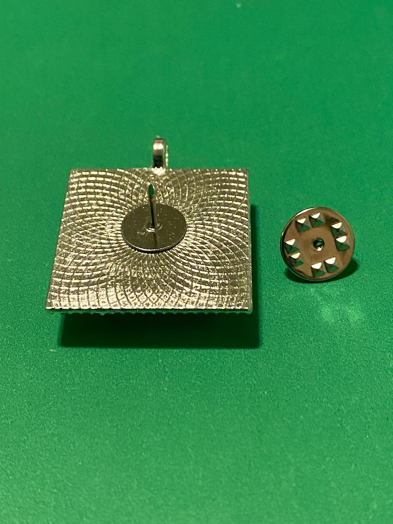 May include: The back of a silver square pin with a decorative pattern. The pin has a silver pin back.