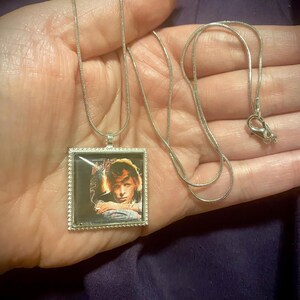 May include: A silver chain necklace with a square pendant featuring a color image of a person with long hair. The pendant has a silver border.