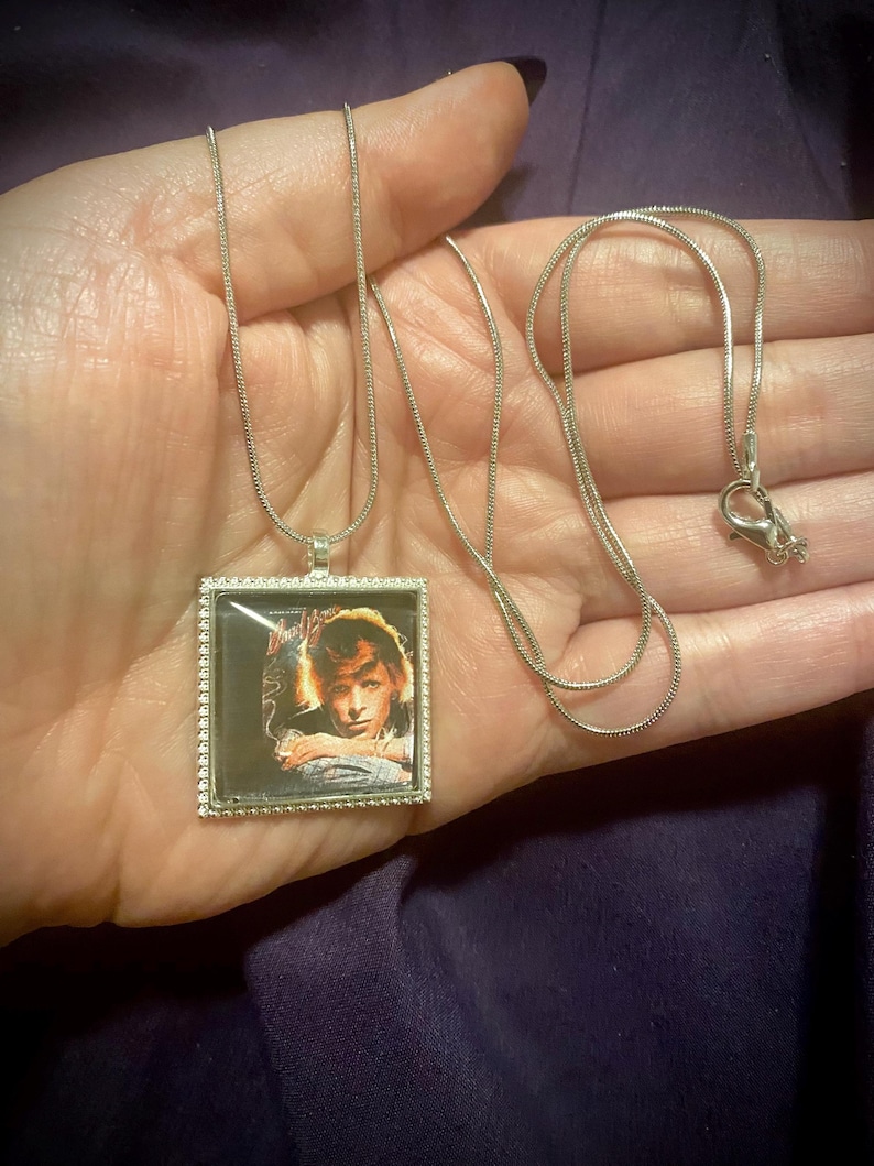 May include: A silver chain necklace with a square pendant featuring a color image of a person with long hair. The pendant has a silver border.