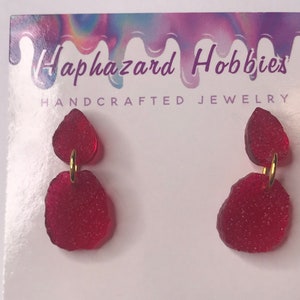 May include: Red, handcrafted earrings with a textured, irregular shape. The earrings are connected by a small gold-colored hoop. The earrings are displayed on a white card with the text "Haphazard Hobbies HANDCRAFTED JEWELRY".
