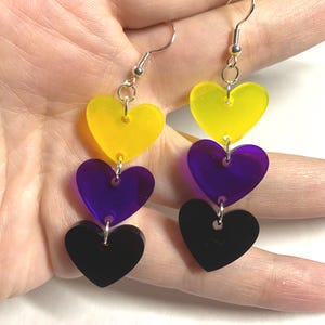 May include: Non-binary pride earrings featuring three heart-shaped charms in yellow, purple, and black. The charms are suspended from silver-colored fish hook ear wires. The earrings are a vibrant accessory.