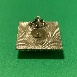 May include: The back of a silver pin with a square design. The pin has a textured surface and a circular pin back.