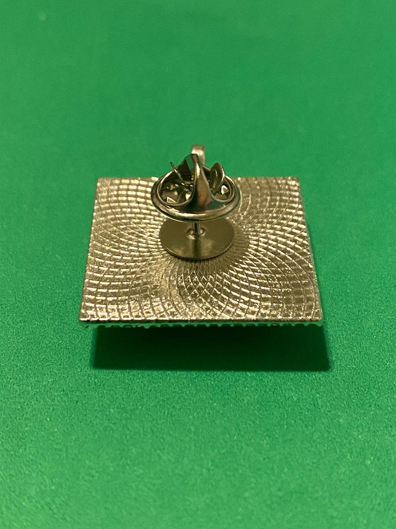 May include: The back of a silver pin with a square design. The pin has a textured surface and a circular pin back.