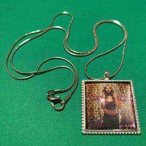 May include: A silver chain necklace with a rectangular pendant featuring a photo of Britney Spears on the cover of her album "...Baby One More Time".