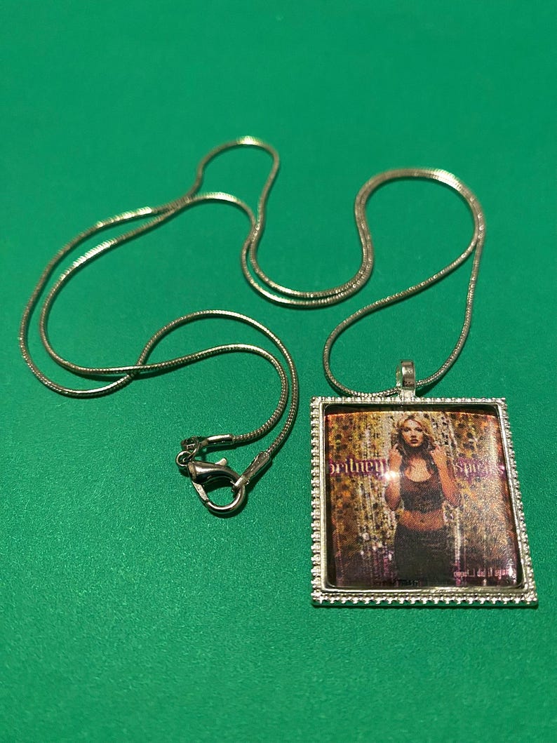 May include: A silver chain necklace with a rectangular pendant featuring a photo of Britney Spears on the cover of her album "...Baby One More Time".