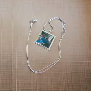 May include: A silver chain necklace with a square pendant featuring a blue and red geometric design. The pendant has a silver border and a clear glass cover.