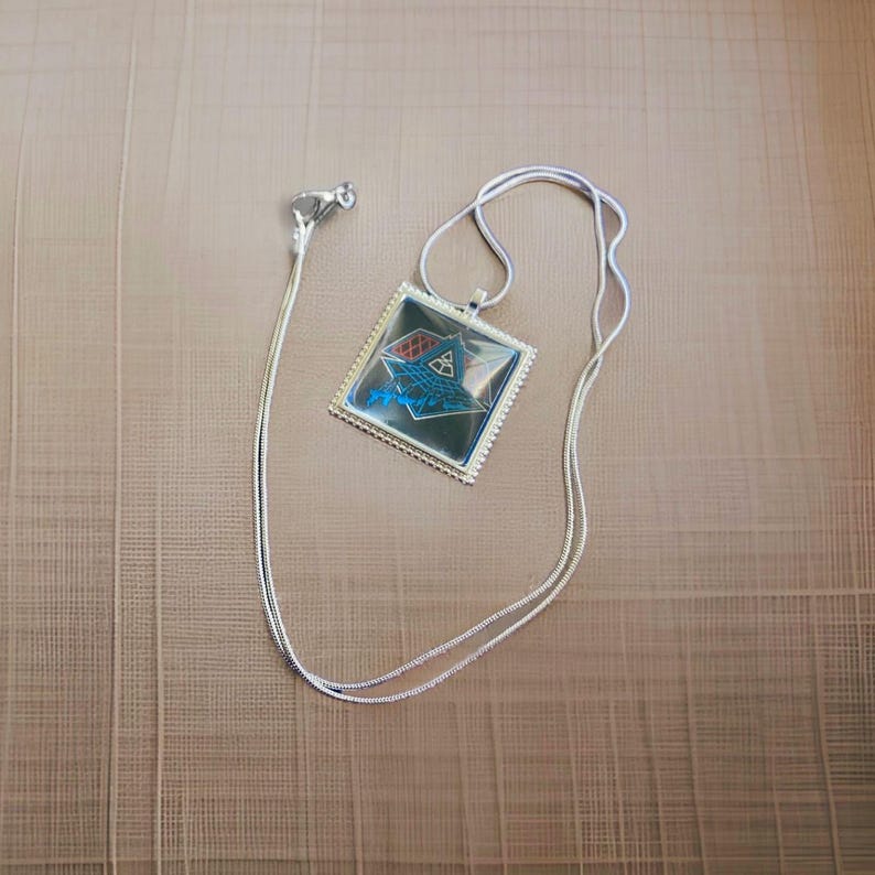 May include: A silver chain necklace with a square pendant featuring a blue and red geometric design. The pendant has a silver border and a clear glass cover.