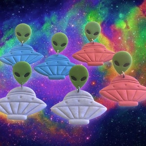 May include: Six pairs of alien-themed earrings. Each earring features a green alien head with large eyes and a flying saucer in blue, white, or pink.