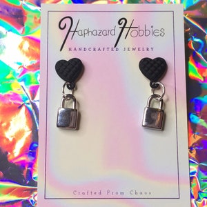 May include: A pair of handcrafted earrings featuring black heart-shaped studs with a quilted texture, connected to silver-colored padlock charms. The earrings are displayed on a card with the text "Haphazard Hobbies HANDCRAFTED JEWELRY Crafted From Chaos."