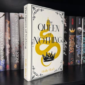 May include: A hardback book titled "The Queen of Nothing" by Holly Black, on a bookshelf. The book features a white cover with black and gold lettering and a snake and crown design. Other books are visible in the background.