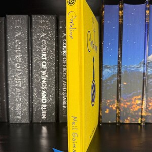 Puede incluir: Un libro de tapa dura de color amarillo brillante titulado Coraline de Neil Gaiman. El lomo presenta un peque&ntilde;o icono de bot&oacute;n en la parte superior, el t&iacute;tulo en letra azul, una silueta de gato azul, un gr&aacute;fico de bot&oacute;n azul y el autor en azul.