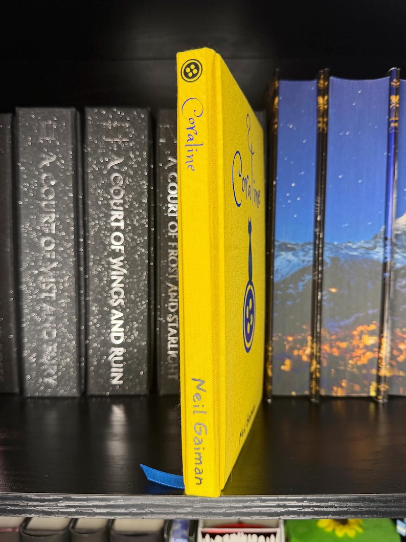 Puede incluir: Un libro de tapa dura de color amarillo brillante titulado Coraline de Neil Gaiman. El lomo presenta un peque&ntilde;o icono de bot&oacute;n en la parte superior, el t&iacute;tulo en letra azul, una silueta de gato azul, un gr&aacute;fico de bot&oacute;n azul y el autor en azul.