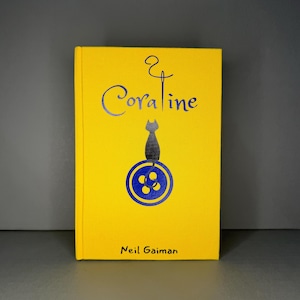 Puede incluir: Un libro de tapa dura amarillo titulado Coraline de Neil Gaiman. La portada presenta un gr&aacute;fico azul oscuro de un gato sentado sobre un bot&oacute;n, con una aguja e hilo formando la letra i en el t&iacute;tulo. El texto est&aacute; impreso en tinta azul oscuro.