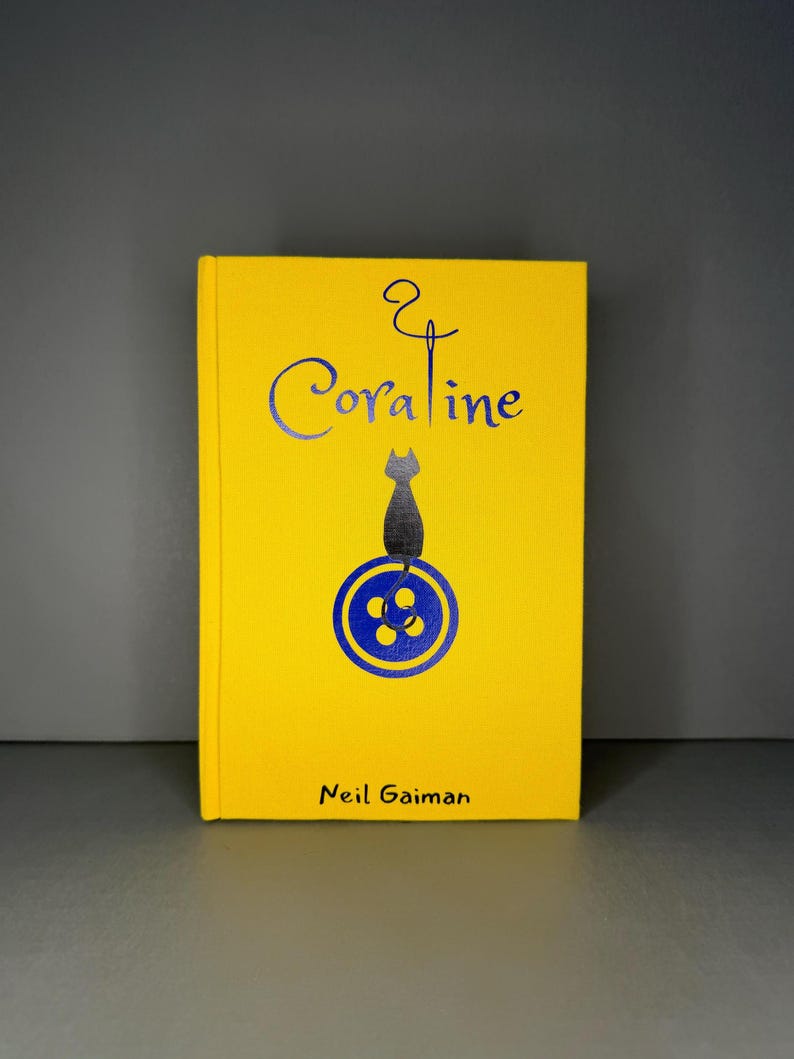 Puede incluir: Un libro de tapa dura amarillo titulado Coraline de Neil Gaiman. La portada presenta un gr&aacute;fico azul oscuro de un gato sentado sobre un bot&oacute;n, con una aguja e hilo formando la letra i en el t&iacute;tulo. El texto est&aacute; impreso en tinta azul oscuro.