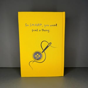 Puede incluir: Una cubierta de libro de color amarillo brillante con un gr&aacute;fico gris oscuro de una aguja de coser enhebrada a trav&eacute;s de un bot&oacute;n de cuatro agujeros. El texto impreso en gris oscuro dice: "So SHARP, you wont feel a thing." Fondo amarillo s&oacute;lido.