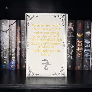 May include: A white book with gold text and decorative black borders, featuring a quote: "She is my wife," and the words "Queen of Elfhame." The book is displayed on a black shelf with other books.