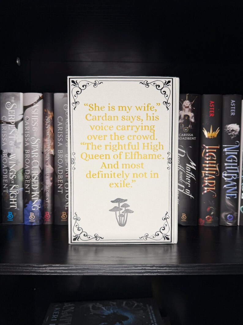 May include: A white book with gold text and decorative black borders, featuring a quote: "She is my wife," and the words "Queen of Elfhame." The book is displayed on a black shelf with other books.