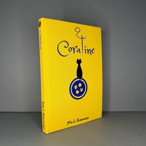 Puede incluir: Un libro de tapa dura de color amarillo brillante titulado Coraline de Neil Gaiman. La portada presenta un gr&aacute;fico azul de aguja e hilo, la silueta de un gato negro y un icono de bot&oacute;n azul. El t&iacute;tulo y el autor est&aacute;n impresos en negro.