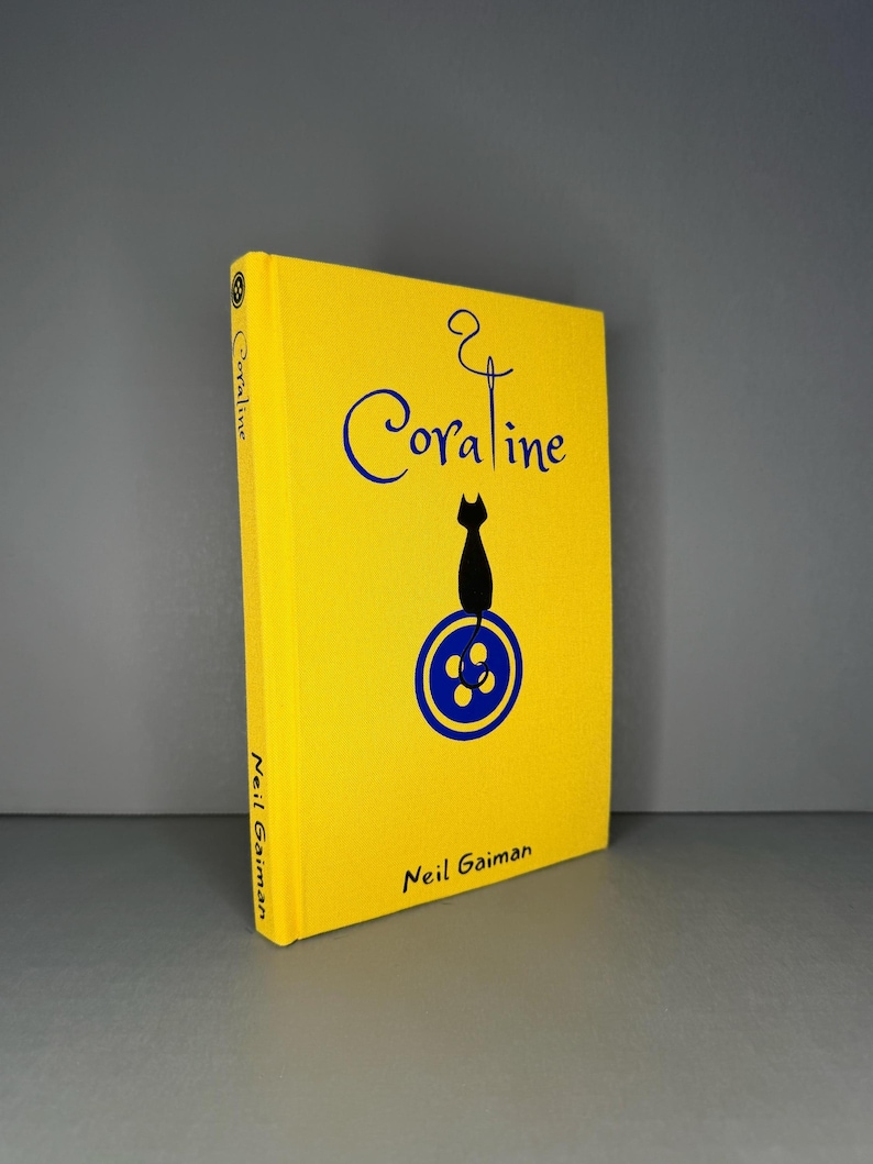 Puede incluir: Un libro de tapa dura de color amarillo brillante titulado Coraline de Neil Gaiman. La portada presenta un gr&aacute;fico azul de aguja e hilo, la silueta de un gato negro y un icono de bot&oacute;n azul. El t&iacute;tulo y el autor est&aacute;n impresos en negro.