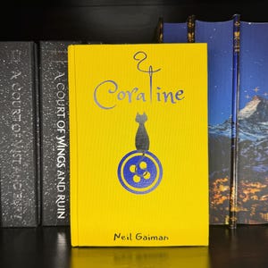 Puede incluir: Un libro de tapa dura titulado Coraline de Neil Gaiman con una cubierta de color amarillo brillante. La portada presenta un gr&aacute;fico azul de un gato sentado sobre un bot&oacute;n, con un dise&ntilde;o de aguja e hilo sobre el t&iacute;tulo. El nombre del autor est&aacute; impreso en negro en la parte inferior.