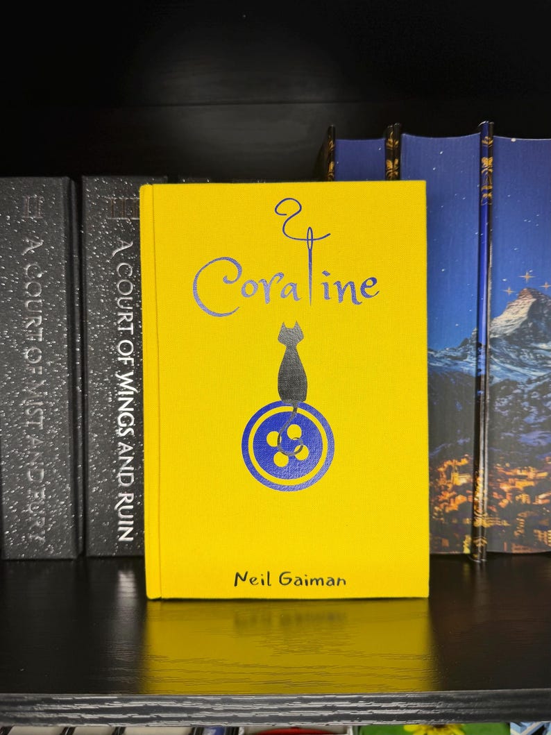 Puede incluir: Un libro de tapa dura titulado Coraline de Neil Gaiman con una cubierta de color amarillo brillante. La portada presenta un gr&aacute;fico azul de un gato sentado sobre un bot&oacute;n, con un dise&ntilde;o de aguja e hilo sobre el t&iacute;tulo. El nombre del autor est&aacute; impreso en negro en la parte inferior.