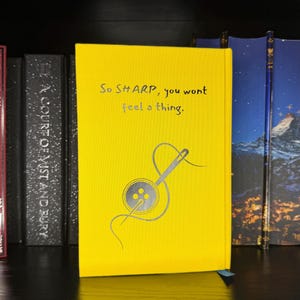 Puede incluir: Un libro de tapa dura de color amarillo brillante con el texto en negro 'So SHARP, you wont feel a thing.' sobre una ilustraci&oacute;n gr&aacute;fica de una aguja de coser enhebrada a trav&eacute;s de un bot&oacute;n de cuatro agujeros, colocado en un estante.