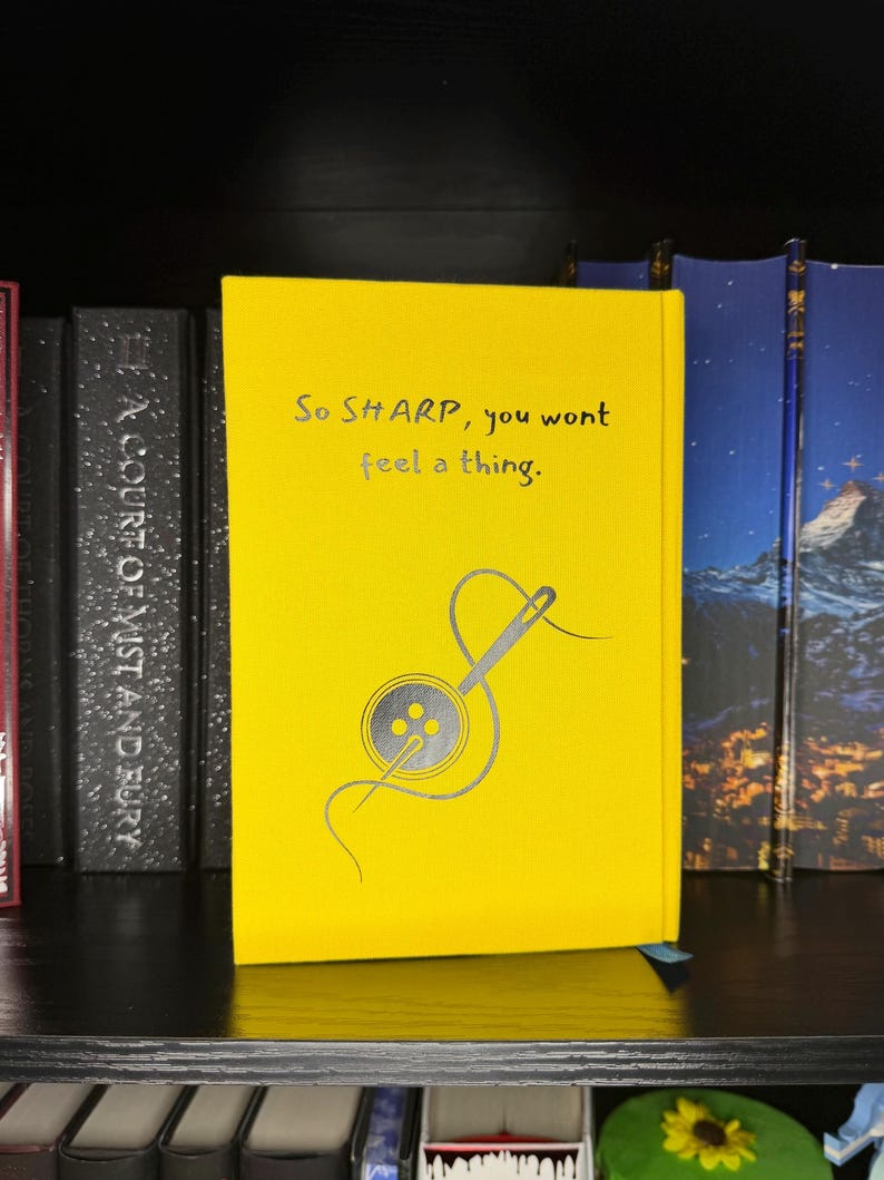 Puede incluir: Un libro de tapa dura de color amarillo brillante con el texto en negro 'So SHARP, you wont feel a thing.' sobre una ilustraci&oacute;n gr&aacute;fica de una aguja de coser enhebrada a trav&eacute;s de un bot&oacute;n de cuatro agujeros, colocado en un estante.