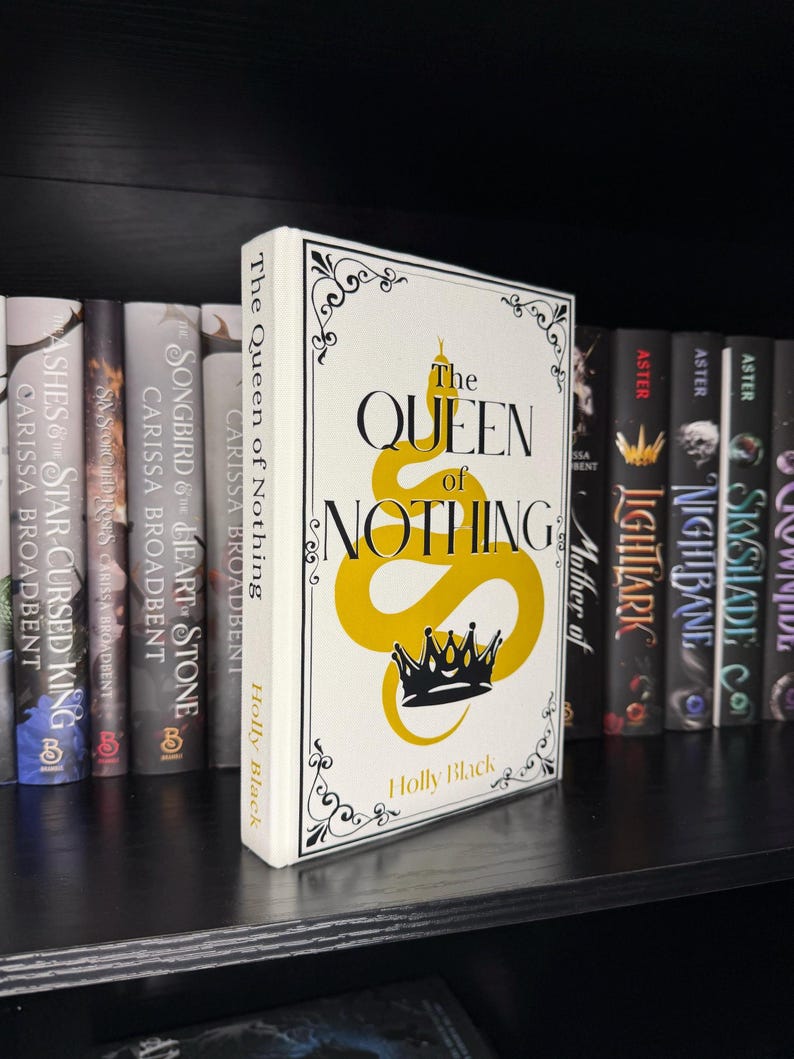 May include: A hardback book titled "The Queen of Nothing" by Holly Black, on a bookshelf. The book features a white cover with black and gold lettering and a snake and crown design. Other books are visible in the background.