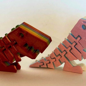 May include: Two 3D printed dinosaur toys, one red with rainbow stripes and one pink with white and blue stripes.
