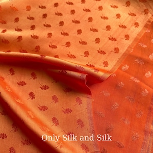 May include: Orange silk fabric with a repeating pattern of small, dark red floral motifs. The fabric is folded, showing the texture and sheen of the material. The text "Only Silk and Silk" is visible.