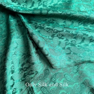 Könnte beinhalten: Smaragdgrüner Seidenstoff mit Paisley-Muster. Der Stoff ist gefaltet, wodurch die Textur und der Glanz des Materials sichtbar werden. Der Text "Only Silk and Silk" ist unten im Bild zu sehen.
