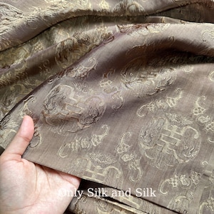 Könnte beinhalten: Nahaufnahme eines braunen Seidenstoffs mit einem sich wiederholenden Muster aus verzierten, stilisierten Blumen- und geometrischen Designs in einer helleren Goldfarbe. Der Stoff hat einen subtilen Glanz und eine luxuriöse Textur. Der Text "Only Silk and Silk" ist sichtbar.