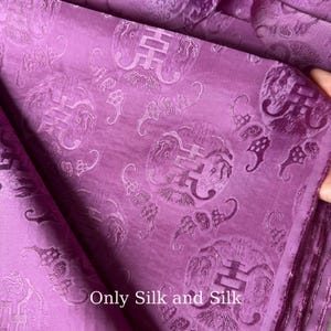 Könnte beinhalten: Nahaufnahme eines lila Seidenstoffs mit einem sich wiederholenden Muster aus verzierten, kreisförmigen Designs und chinesischen Schriftzeichen. Der Stoff hat einen dezenten Glanz und der Text "Only Silk and Silk" ist unten sichtbar.