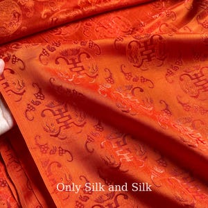 Può includere: Primo piano di un vibrante tessuto di seta arancione con un motivo ripetuto di simboli stilizzati e motivi floreali. Il tessuto ha una lucentezza sottile ed è piegato, rivelando la sua consistenza. Il testo in basso recita "Only Silk and Silk."